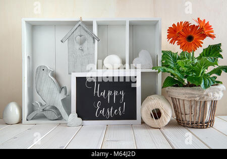 Frühling Hintergrund mit Frühling Dekorationen. Vitrine mit verschiedenen Objekten und Orange Gerbera Flower Pot. Gerne Worte "Ostern" auf dem Schwarzen Stockfoto