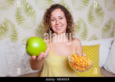 Junge lockige Frau, Sitzen, Sofa und ein grüner Apfel und eine Vase mit Chips. Gesunde Ernährung Konzept. Stockfoto