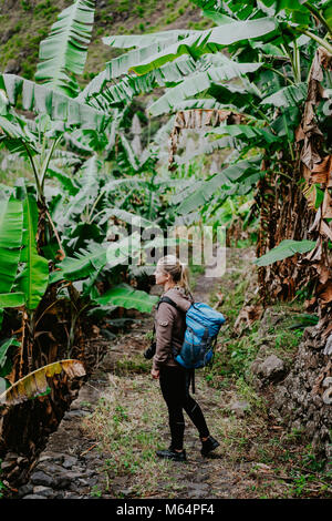 Blond yong Frauen mit Kamera und blau Rucksack zu Fuß durch Bananenplantagen auf der trekkingroute Tal zu Paul. Santo Antao Insel. Kap Verde Stockfoto