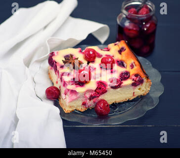 Stück Käsekuchen mit Kirschen Beeren auf einer Glasplatte, hinter ein Glas eingemachte Kirschen Stockfoto