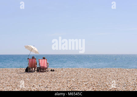 Die steigenden Temperaturen an der Südküste von England als Paar entspannen Sie sich auf traditionelle gestreifte Liegestühle mit Sonnenschirm auf einem glühend heißen Tag in Lyme Regis. Stockfoto