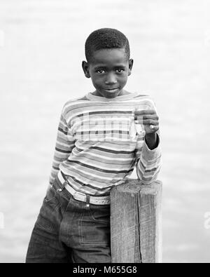 1970 s BAHAMIAN JUNGEN im gestreiften Hemd mit einem traurigen Gesichtsausdruck in die Kamera auf der Station, DIE BIS ZWEI SEHR KLEINE FISCHE-a8177HAR 001 HARS NOSTALGIE SEHR AUGENKONTAKT 10-12 JAHRE 7-9 JAHRE JUGENDLICHE JUNGE GRÖSSE GLÜCK ZWEI TIERE BAHAMAS afrikanische Amerikaner, AFRICAN-AMERICAN, FREIZEIT SCHWARZ ETHNIZITÄT afrikanische Amerikaner stolz AFRICAN AMERICAN LÄCHELT FRÖHLICH KLEINE JUNGEN MÄNNER vor - jugendlich PRE-TEEN BOY B&W BAHAMIAN SCHWARZ UND WEISS AUF KAMERA ALTMODISCH Stockfoto