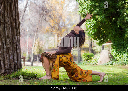 Reife Frau übt Yoga im Park an einem sonnigen Tag. Tragen bunte Hosen und braun Shirt auf Krieger aufwerfen, wie virabhadrasana bekannt Stockfoto