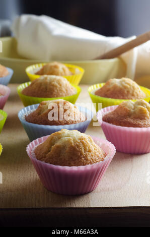 Reihen der frisch gebackene cup cakes in bunten Fällen auf Holz Schneidebrett in der sonnendurchfluteten Küche. Schüssel und Löffel aus Holz im Hintergrund. Stockfoto