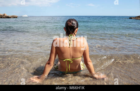 Rückansicht der Frau, die im flachen Wasser am Strand, Punta Negra, Mallorca, Spanien sitzt Stockfoto