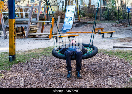 Berlin, Mitte, Senior älterer Mann entspannt auf Schwingen des Kindes in der Kinder Spielplatz Stockfoto