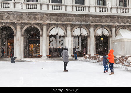 Venedig, Venetien, Italien, 1. März 2018. Schlechtes Wetter in Venedig heute mit Sub-zero temperatues zwischen minus 3 und minus 2 und kontinuierliche Schnee den ganzen Tag durch das Tier aus dem Osten, oder die Sibirische Front aus Russland quer durch Europa verursacht. Frau und ihre beiden Kinder außerhalb Florians in der Piazza San Marco. Wie die Kinder spielen Sie schaut sehnsüchtig auf das warme Interieur des Restaurants. Stockfoto