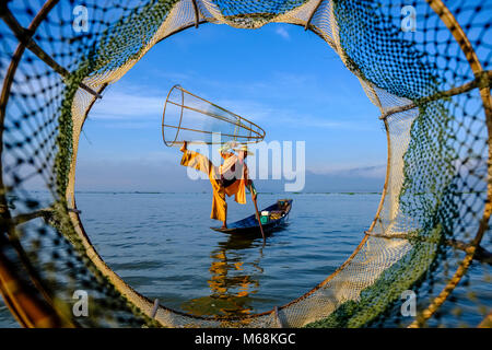 Ein Fischer, stehend auf seinem Boot, posierend, ist die traditionelle Art und Weise auf dem Inle See. Gesehen durch eine basketlike Fischernetz Stockfoto