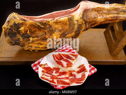 Nahaufnahme von Serrano Schinken Bein mit Platte und Tischdecke über Schwarz Stockfoto