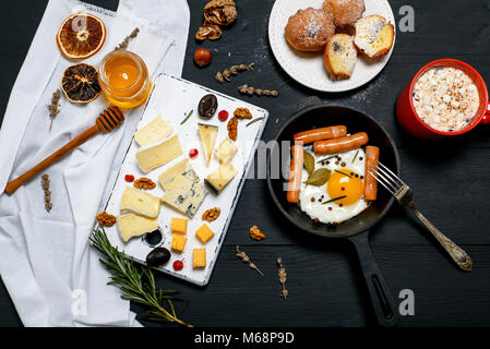 Spiegelei mit Würstchen in einem schwarzen runden gusseisernen Pfanne, rot Tasse mit Kakao und Marshmallow- und Käseplatte mit verschiedenen Sorten Käse Stockfoto