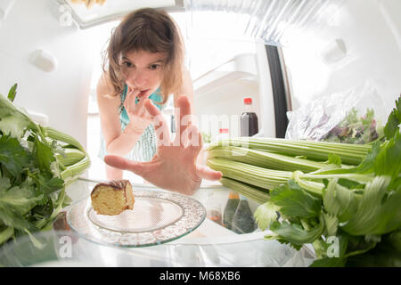 Ernährung Kampf: Eine Hand packte einen kleinen Stück Süßes aus der offenen Kühlschrank voller Grüns. Stockfoto