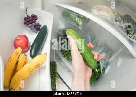 Gesunde Ernährung: Eine Hand packte eine Gurke aus dem offenen Kühlschrank voller Grüns. Stockfoto