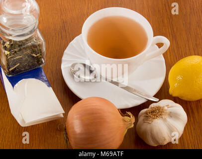 Eine Tasse Tee mit Untertasse und Löffel, Zitrone, Zwiebel, Knoblauch, ein Glas Dosis mit Kaffee, Papiertaschentücher. Alles, was gegen die Kälte auf dem hölzernen de Fangen Stockfoto