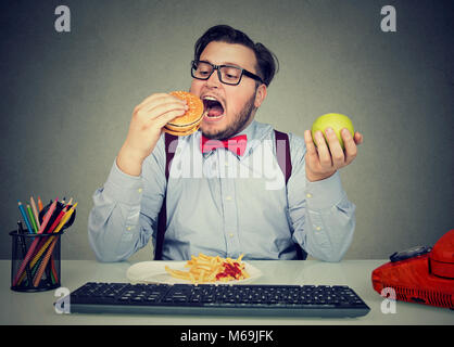 Junger Mann essen Hamburger mit Pommes anstelle von Apple in Bewegungsmangel im Büro. Stockfoto