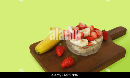 Glasplatte mit gehackt Erdbeeren und Bananen Stücke auf einem dunklen Holzplatte mit 3 Erdbeeren und eine Banane mit Schale auf einem grünen Zurück eingerichtet Stockfoto