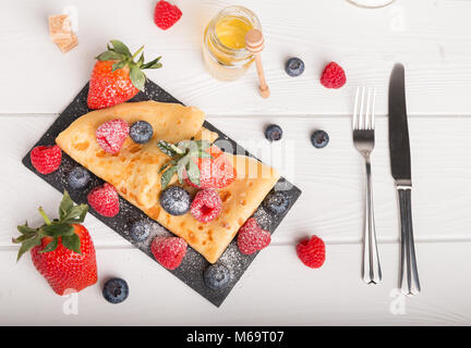 Pfannkuchen Teller mit frischem Obst Erdbeere Heidelbeere und Himbeere auf einem weißen Holztisch. Auf der Seite Kaffee und Honig und Zucker. Stockfoto