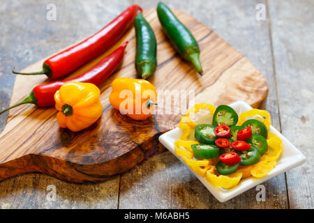 Rote, grüne und gelbe Peperoni auf Holzbrett mit in Scheiben geschnittenen Paprika in weiße Schüssel auf alten Holztisch Stockfoto