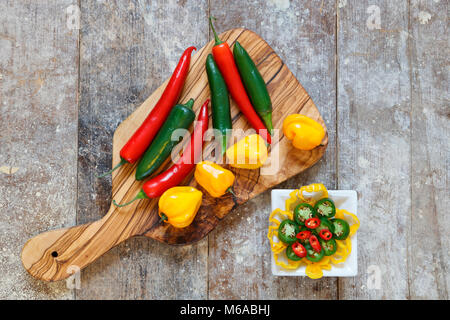 Ansicht von oben der rote, grüne und gelbe Peperoni auf Holzbrett mit in Scheiben geschnittenen Paprika in weiße Schüssel auf alten Holztisch Stockfoto