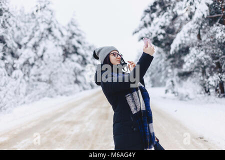 Ein hübsches Mädchen macht einen selfie mitten in einem verschneiten Wald Straße im Winter Stockfoto