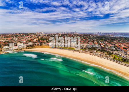 Surfer auf Emerald Wellen der berühmte Bondi Beach in Australien, Sydney, mit weit entfernten Stadt CBD Türme am Horizont und blauen bewölkten Himmel von Sommer. Stockfoto