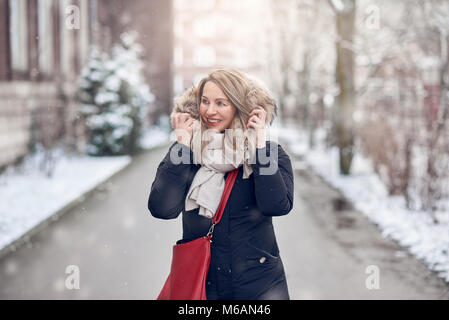 Lächelnden jungen Frau entlang eine verschneite Straße im Winter halten der Pelzbesatz auf ihre Jacke auf der Seite mit einem glücklichen Lächeln Stockfoto