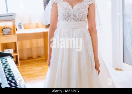 Braut im weißen Kleid neben Klavier im Wohnzimmer. Fokus auf Kleid! Stockfoto