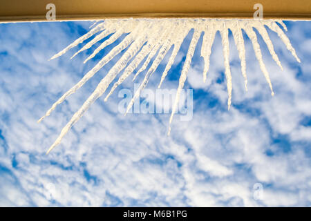 Closeup Halbkreis von großen funkelnden Eiszapfen an Fensterrahmen gegen hellen blauen bewölkten Himmel Stockfoto