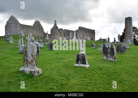 Verwitterte Grabsteine mit Ruinen der mittelalterlichen Kirchen und runder Turm im Hintergrund, im Kloster Clonmacnoise in Irland. Stockfoto