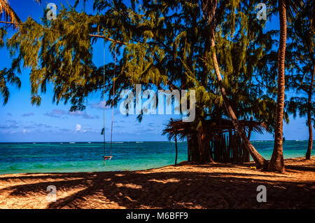 Eine Holzhütte auf dem Sand und eine Schaukel im Schatten der Bäume und Palmen am Ufer des Blauen Ozean mit weißen Wellen, Himmel und Wolken o Stockfoto