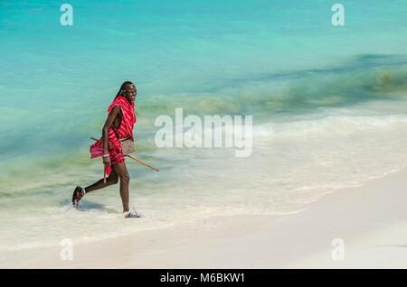 Sansibar, Tansania - November 30, 2014: junge massai Mann mit farbenfrohen traditionellen Kleidung und Kopfbedeckung zu Fuß durch das Wasser auf der Nungwi Beach Stockfoto