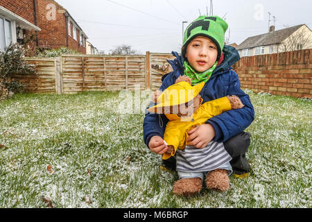 Ein Junge spielt mit seinem Teddybären im vorderen Garten im Schnee. Sowohl das Kind als auch der Spielzeug sind im Winter Hüte und Mäntel gekleidet. Stockfoto