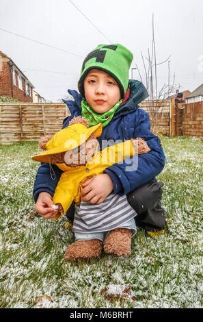 Ein Junge spielt mit seinem Teddybären im vorderen Garten im Schnee. Sowohl das Kind als auch der Spielzeug sind im Winter Hüte und Mäntel gekleidet. Stockfoto