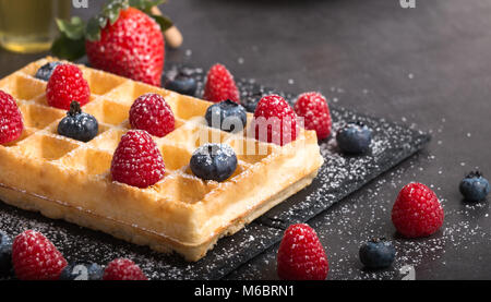 Frisch gebackene Brüsseler Waffeln auf einem schwarzen Stein Teller mit frischen Erdbeeren und Heidelbeeren und Himbeeren auf einem dunklen Stein Tabelle mit Honig und Cof Stockfoto