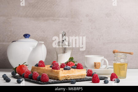 Frisch gebackene Brüsseler Waffeln auf einem schwarzen Stein Teller mit frischen Erdbeeren und Heidelbeeren und Himbeeren auf einem weißen Tisch mit Honig und c Stockfoto