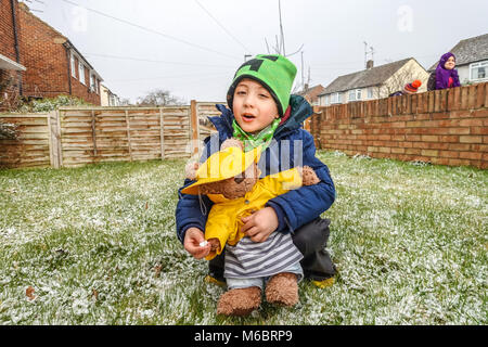 Ein Junge spielt mit seinem Teddybären im vorderen Garten im Schnee. Sowohl das Kind als auch der Spielzeug sind im Winter Hüte und Mäntel gekleidet. Stockfoto