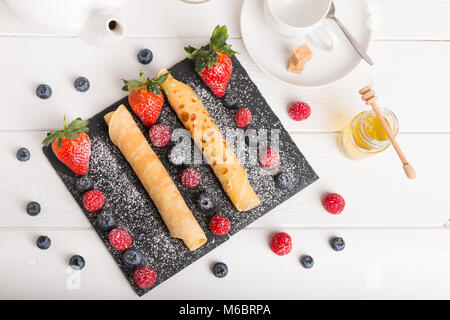Gerollte Pfannkuchen mit frischen Erdbeeren und Himbeeren und Blaubeeren auf einen schwarzen Stein Platte auf eine weiße Holztisch mit Honig und weißer Zucker und Stockfoto