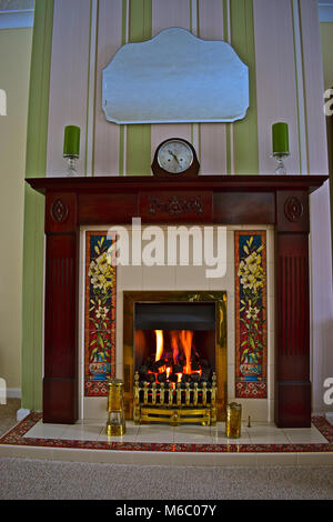 Ein gemütliches Bild eines Kohle- wirkung Gas Feuer mit Holz Kaminsims oben mit einem 50er Ära Uhr durch zwei Kerzen flankiert. Messing Miners Lamp unten. Stockfoto