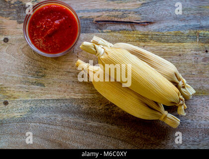 Drei ganze, wickelte Tamales auf Holztisch mit Hot Tomato Sauce im Glas serviert, und viele Kopieren - Raum, von oben gesehen - Mexikanische Foo Stockfoto
