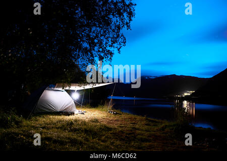 Camping mit einem beleuchteten Zelt am Strand und Fjord in den Bergen von Norwegen während der Nacht mit einem bewölkten Himmel und Sterne. Stockfoto