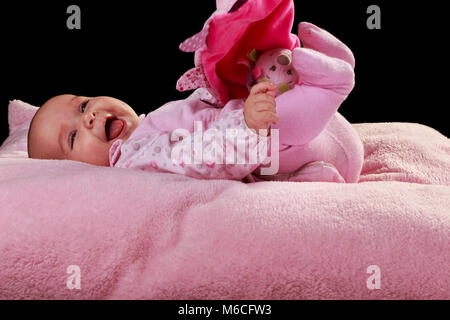 Fröhliches Baby Mädchen liegt auf einem rosa Kissen Spielzeug spielen mit einem weichen Tuch. auf schwarzem Hintergrund Stockfoto