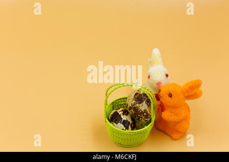 Zwei bunte Osterhasen und einem grünen Korb mit Eiern auf orangem Hintergrund, Ostern Dekoration. horizontale Ansicht. Stockfoto