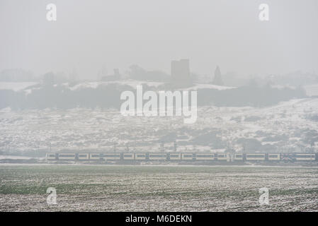 Der C2C-Eisenbahnzug führt durch den schneebedeckten Hadleigh Country Park und die Felder der Hadleigh Marshes mit Ruinen von Hadleigh Castle an nebelig winterlichen Tagen während des östlichen Wetterphänomens. Essex ländlich Stockfoto