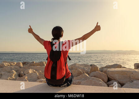 Junge attraktive Frau, Daumen oben auf den Händen zu sitzen, der am Ufer des Meeres. Stockfoto