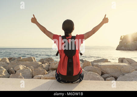 Junge attraktive Frau, Daumen oben auf den Händen zu sitzen, der am Ufer des Meeres. Stockfoto