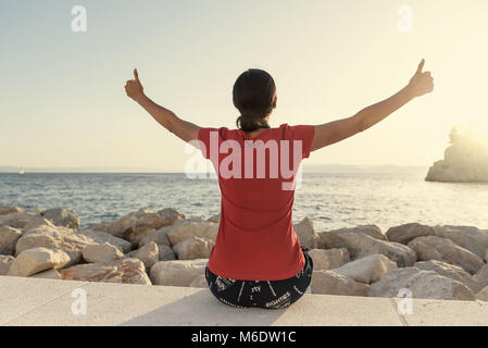 Junge attraktive Frau, Daumen oben auf den Händen zu sitzen, der am Ufer des Meeres. Stockfoto
