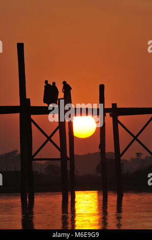 Die U-Bein Brücke aus Teakholz in Amarapura Myanmar bei Sonnenuntergang vom Wasser aus gesehen mit drei Mönche gehen, um das Wasser zu überqueren Stockfoto