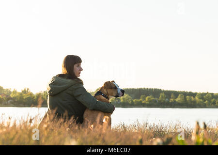 Hundehalter und Ihr Haustier sitzen am Flussufer bei Sonnenuntergang Stockfoto