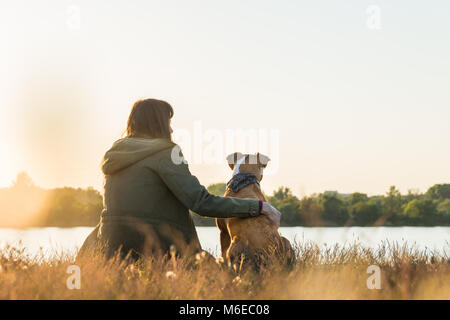 Hundehalter und Ihr Haustier sitzen am Flussufer bei Sonnenuntergang Stockfoto