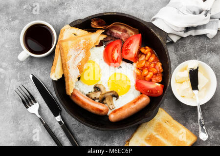 Traditionelles Englisches Frühstück - Speck, Würstchen, Spiegeleier, Tomaten, Bohnen, Pilze, Toast mit Butter - beim Kochen, Pan und Kaffee auf einem grauen zurück Stockfoto