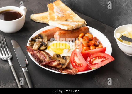Traditionelles Englisches Frühstück - Speck, Würstchen, Spiegeleier, Tomaten, Bohnen, Pilze, Toast mit Butter - In weißen Teller und Kaffee auf einem dunklen zurück Stockfoto